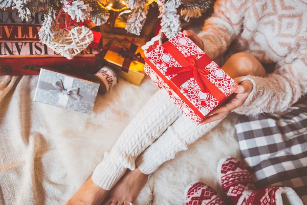 Fiatal, csinos nő a meleg és kényelmes ruhát ül a könnyű fa padló világos házát a karácsonyfa közelében, és ajándék doboz tart a kezében. — Stock Fotó
