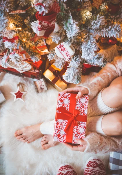 Junge hübsche Frau in warmen, gemütlichen Kleidern sitzt auf dem hellen Holzboden in ihrem hellen Haus neben dem Weihnachtsbaum und hält eine Geschenkschachtel in der Hand. — Stockfoto