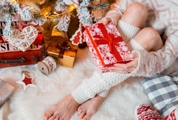 Jeune jolie femme dans des vêtements chauds et confortables est assis sur le sol en bois clair dans sa maison lumineuse près de l'arbre de Noël et tient boîte cadeau dans ses mains . — Photo