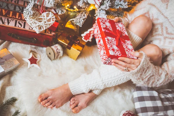 Junge hübsche Frau in warmen, gemütlichen Kleidern sitzt auf dem hellen Holzboden in ihrem hellen Haus neben dem Weihnachtsbaum und hält eine Geschenkschachtel in der Hand. — Stockfoto