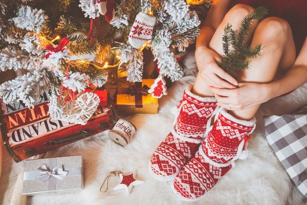 Mladá krásná žena v teplé příjemné oblečení sedí na lehké dřevěné podlaze v jejím světle domě poblíž vánoční strom a drží krabičky ve svých rukou. — Stock fotografie