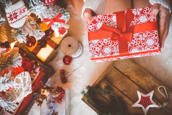 Jonge mooie vrouw in warme gezellige kleren zit op de lichte houten vloer in haar heldere huis in de buurt van de kerstboom en houdt van luxe-geschenketui in haar handen. — Stockfoto