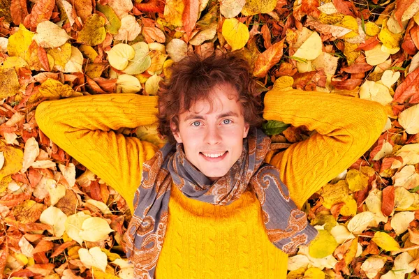 Gekrulde man in de gele trui en sjaal liggen op Herfstbladeren가 잎에 노란 스웨터와 스카프 거짓말 곱슬 남자 — Stockfoto