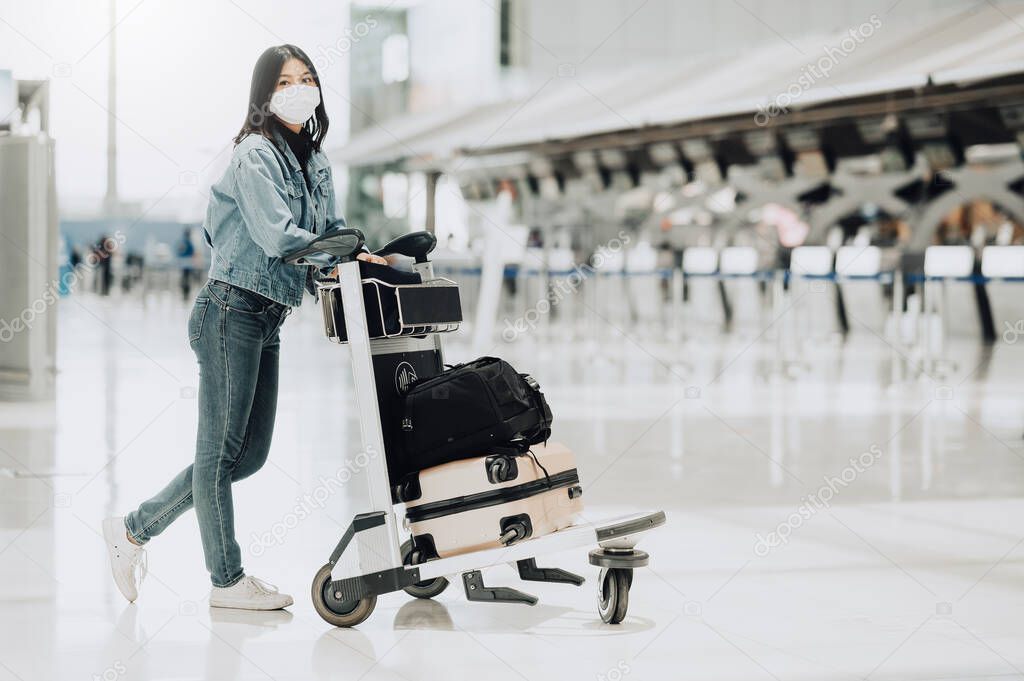 Happy Asian tourist woman with mask protection for coronavirus walking with luggage trolley ready for travel