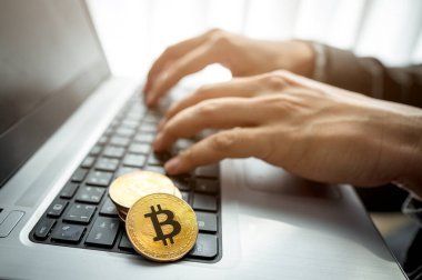 Not defterinde Bitcoin sembolü olan bir yığın altın sikke ve arka planda dizüstü bilgisayar kullanan bir işadamı eli. İşadamı kripto para ticareti yapıyor