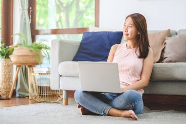 Güzel Asyalı kadın dizüstü bilgisayarını kullanırken gülümsüyor. Oturma odasında yerde oturuyor.