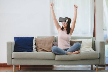 Güzel, mutlu, heyecanlı, VR kulaklıklı Asyalı kadın evde sanal gerçeklikte başarı ve zaferi kutlamak için iki elini kaldırıyor.