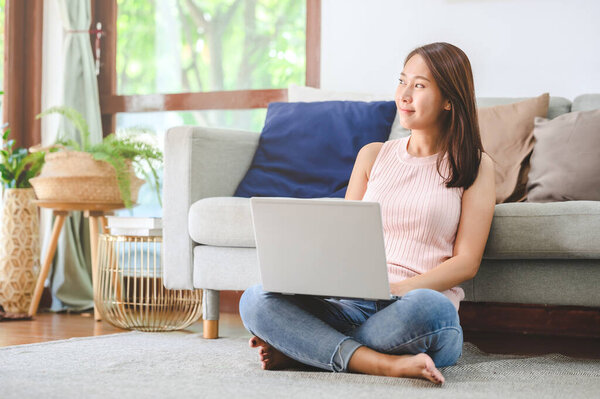 Красивая азиатка улыбается при использовании ноутбука, сидя на полу в гостиной дома