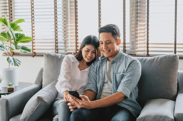 Happy Asian couple using smartphone mobile on sofa at home.