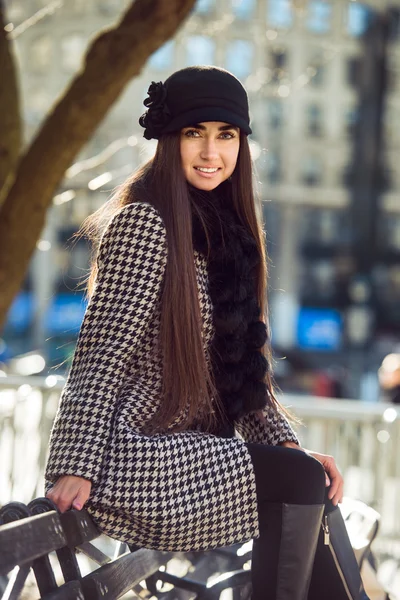 Mulher feliz bonita sentada no banco no dia ensolarado primavera vestindo casaco e chapéu da moda — Fotografia de Stock