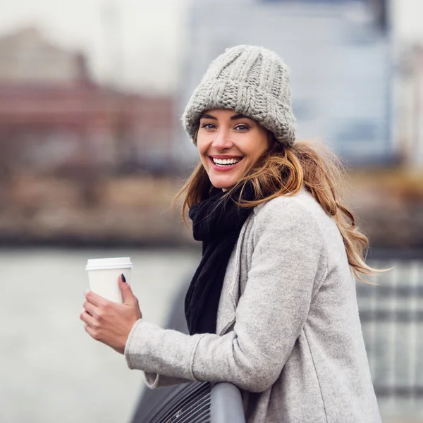 Beautiful smiling woman drinking coffee from paper cup standing on river embankment and enjoing the view — Stock Photo, Image