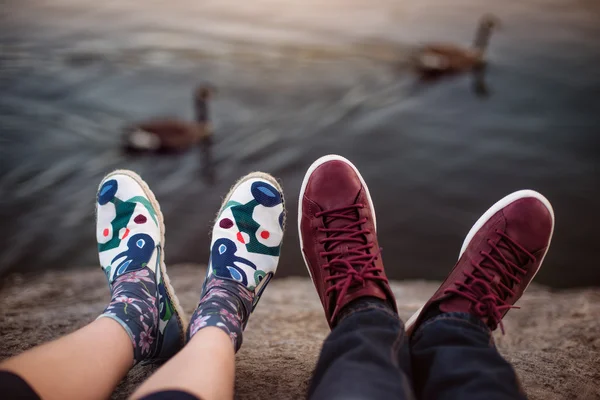 Los pies con los zapatos de la pareja en la cita romántica sentado en las rocas cerca del lago —  Fotos de Stock