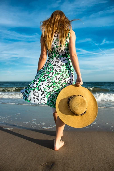 Mujer joven sosteniendo sombrero y caminando en la arena de la playa hasta el océano. Foto de la mujer de atrás —  Fotos de Stock