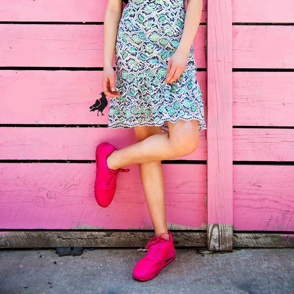 Tênis rosa em pernas de menina no fundo da parede rosa de madeira grunge. Foto de rua. Menina vestindo tênis e saia de verão — Fotografia de Stock