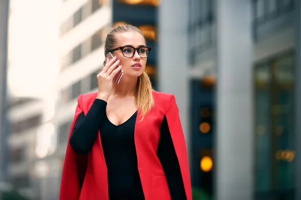 Κομψό επιχειρηματίας με τα πόδια στην οικονομική περιοχή της πόλης και μιλάμε για κινητό τηλέφωνο φορώντας μπουφάν και γυαλιά. Επιτυχημένη γυναίκα των επιχειρήσεων το περπάτημα κοντά ουρανοξύστες. — Φωτογραφία Αρχείου