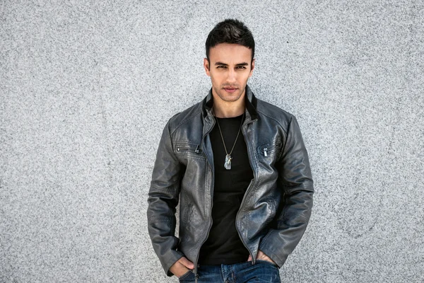 Krásný módní člověk pózuje u zdi na sobě šedý kožený kabát, černé tričko a džíny. — Stock fotografie