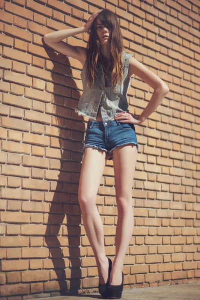 Linda jovem morena em jeans roupas posando ao ar livre perto de parede de tijolo vermelho — Fotografia de Stock
