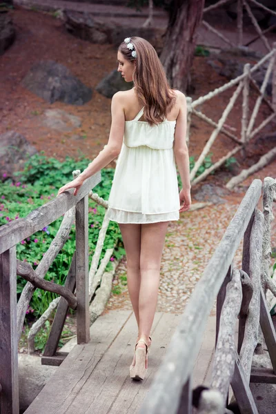 Mulher bonita com pernas longas vestindo vestido branco andando na ponte em uma floresta — Fotografia de Stock