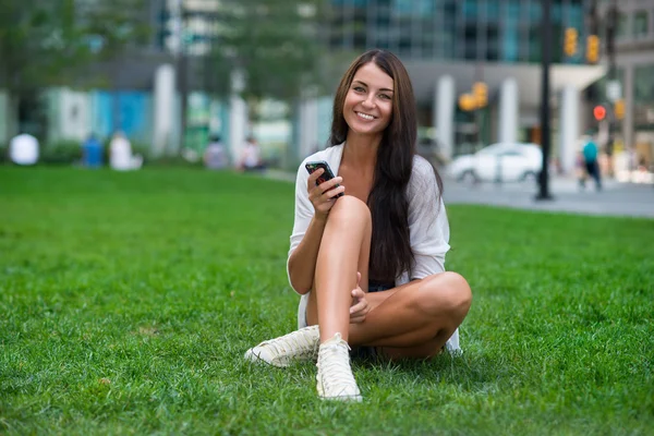 Beautiful woman sitting on the green grass at city park and showing victory v hand sign — Stock Photo, Image