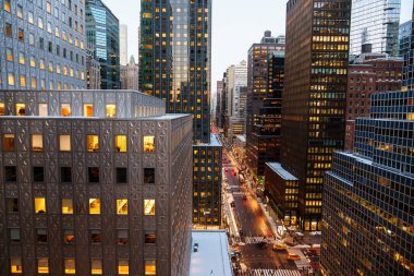 Manhattan, New York 'un yüksek açılı manzarası altında yoğun bir cadde oluşturan modern gökdelenlerin yoğun bir kümesini gösteriyor. Her iki taraftan da uzun cam ve çelik ofis kuleleri yükseliyor. Işıklı pencereleri sıcacık parlıyor.