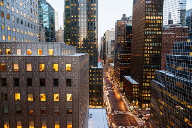 Manhattan, New York 'un yüksek açılı manzarası altında yoğun bir cadde oluşturan modern gökdelenlerin yoğun bir kümesini gösteriyor. Her iki taraftan da uzun cam ve çelik ofis kuleleri yükseliyor. Işıklı pencereleri sıcacık parlıyor.