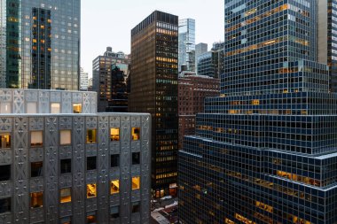 Manhattan, New York 'un yüksek açılı manzarası altında yoğun bir cadde oluşturan modern gökdelenlerin yoğun bir kümesini gösteriyor. Her iki taraftan da uzun cam ve çelik ofis kuleleri yükseliyor. Işıklı pencereleri sıcacık parlıyor.