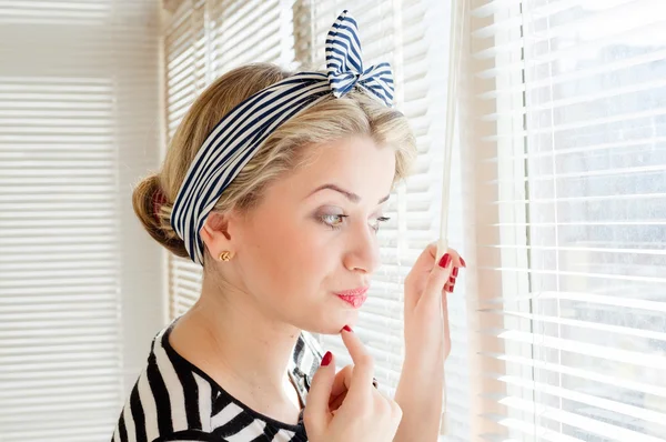 Beautiful blond pinup woman looking thoughtfully through jalousie windows — Stock Photo, Image