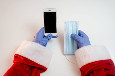 Tıbbi maskeli akıllı telefonlu Noel Baba. İnternetten selamlar, Noel ve yeni yıl için servis siparişleri. Koronavirüs salgınında sosyal mesafe, yeni bir gerçeklik..
