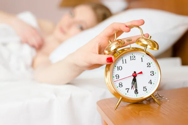 Beautiful blond young woman blocking alarmclock off wake up time & looking at clock — Stock Photo, Image