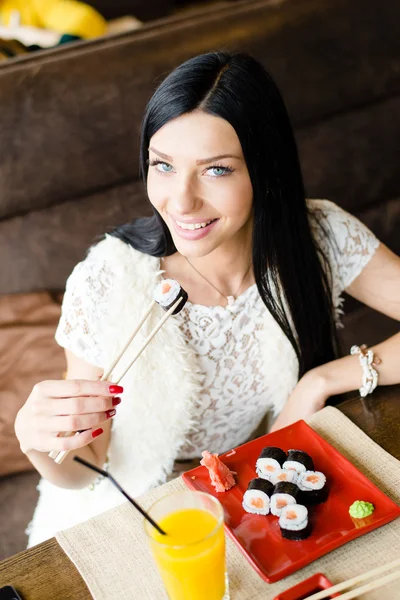 Usmívající se sushi: detailní obrázek nádherná bruneta mladá žena jíst chutné japonské sushi s zábava sedí & šťastný při pohledu na fotoaparát na pozadí interiéru restaurace — Stock fotografie