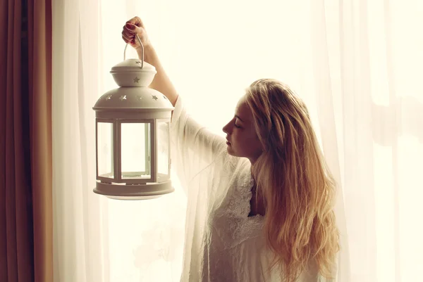 Imagen de la hermosa mujer joven rubia sosteniendo la antorcha de la luz de la vela en la noche temprana y mirando el espacio de copia en el fondo de la ventana de luz —  Fotos de Stock
