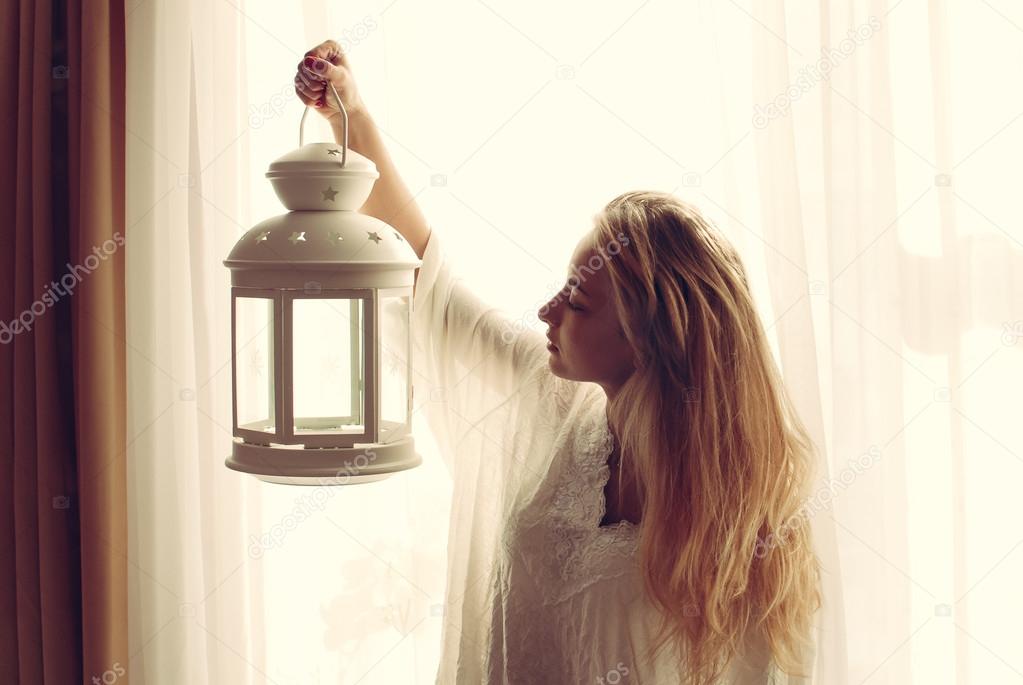 image of beautiful blond young woman holding candle light torch in the early evening & looking at copy space on light window background