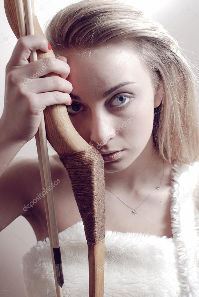 Picture of amazon warrior girl beautiful blond young woman holding bow & arrow close to herself & looking at camera on light wall background closeup portrait