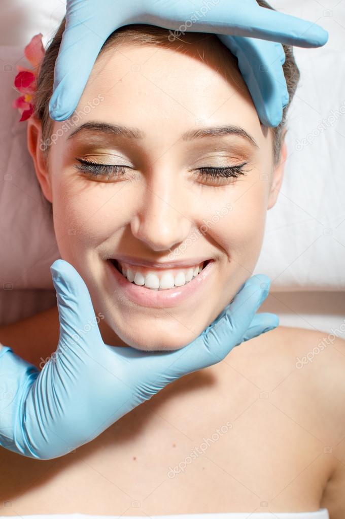 Enjoyable face massage: close up of beautiful young woman sexy attractive girl with great dental care having fun on spa procedures happy smiling relaxing eyes closed & laying on white bed portrait