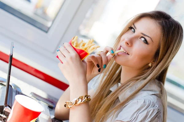 Working lunch: close up image of drinking & eating fries beautiful young business woman blond girl having fun working on laptop pc computer in restaurant or coffee shop smiling & looking at camera — Stock Photo, Image