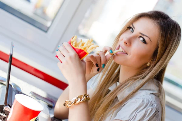 Roboczy lunch: bliska obraz picia & jedzenia frytek piękny młody biznes blond dama dziewczynka zabawy pracy na komputerze pc laptopa w restauracji lub kawiarni uśmiechający się & patrząc na kamery — Zdjęcie stockowe