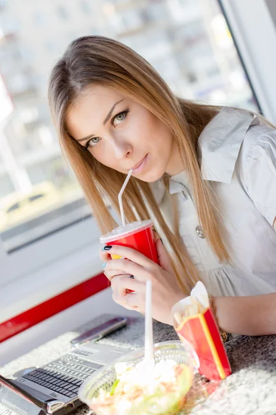 Comer trabajando: primer plano retrato de beber y comer hermosa joven mujer de negocios linda chica rubia divertirse trabajando en el ordenador portátil PC en el restaurante o cafetería sonriendo y mirando a la cámara — Foto de Stock