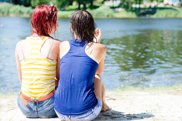 2 lindas novias mujeres jóvenes en camisas de ropa mojada que se divierten relajándose sentado en la orilla del río en el suelo de playa de arena y hablando juntos en verano verde al aire libre copiar el espacio de fondo — Foto de Stock