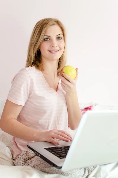 Vacker mild söt ung kvinna blå ögon blond tjej i säng med laptop pc dator äta äpple och tittar upp på lätt kopiera utrymme bakgrund närbild porträttbild — Stockfoto