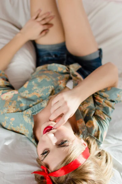 Young beautiful blond pinup girl in jeans shorts flower print shirt having fun relaxing in white bed lying on back putting her feet on wall happy smiling & looking up at copy space portrait image — Stock Photo, Image