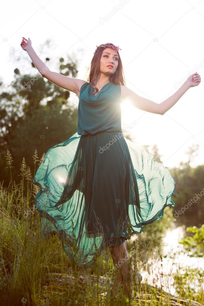 image of young woman beautiful brunette girl having fun standing in grass in green dress with pink wreath of flowers with wide-open arms against sun light & looking at outdoors copy space background