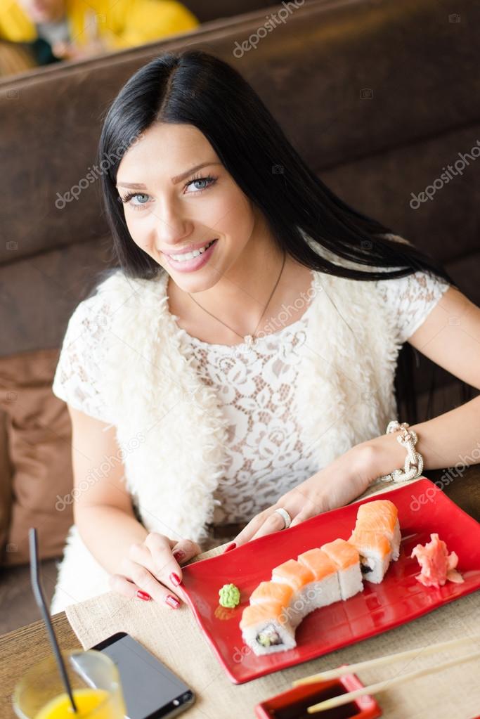 Young beautiful girl blue eyes sexy female sitting in a restaurant or coffee shop with a plate of sushi, orange juice and mobile telephone having fun happy smiling & looking at camera portrait