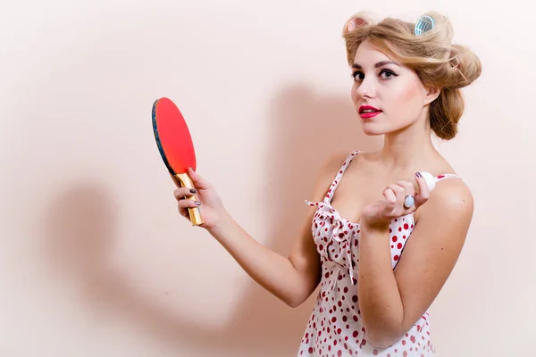 Portrait of elegant attractive glamor pinup girl dazedly looking at camera posing in red dress with flower in hair & holding bat ball for table tennis on white or light copy space background closeup — Stock Photo, Image