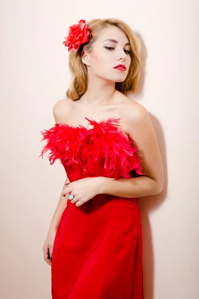 Fashion glamor young blond pin up lady having fun sensually dreaming in red dress with feathers & open naked shoulders, flower in hair posing over white or light copy space portrait picture — стоковое фото