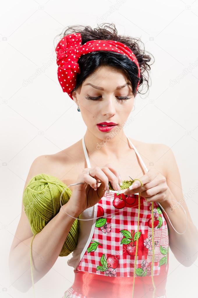 close up on sexy funny young brunette pin up lady having fun wearing apron and red bow learning how to knit over white or light copy space background portrait picture