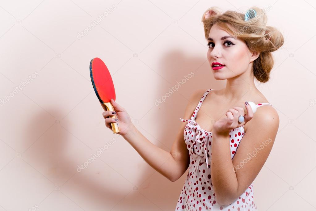 Portrait of elegant attractive glamor pinup girl dazedly looking at camera posing in red dress with flower in hair & holding bat ball for table tennis on white or light copy space background closeup