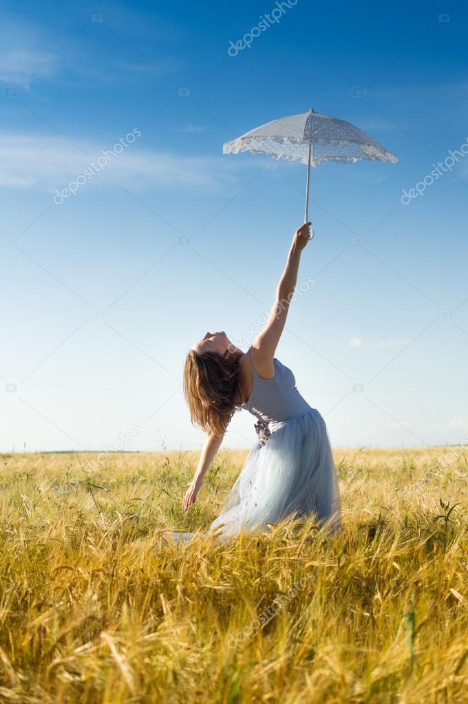 alone in the field: elegant romantic beautiful young lady in long white dress having fun holding up parasol standing back to camera on blue sky copy space background