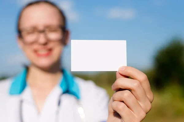 Close-up op visitekaartje: vrouwelijke arts houden en lege kaart gelukkig lachend & camera kijken op blauwe hemel kopie ruimte achtergrond buiten tonen — Stockfoto