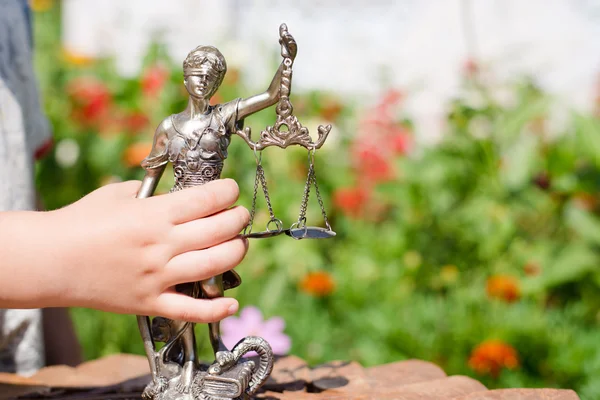 Mano de niño sosteniendo escultura de ellos, femida o diosa de la justicia sobre hojas verdes fondo bokeh natural — Foto de Stock