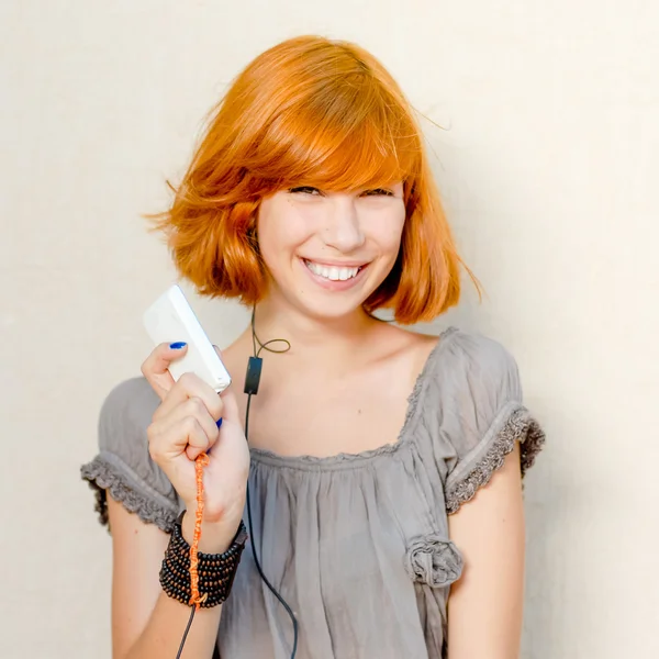 Portrait of happy smiling beautiful young woman with red hair having fun holding mobile digital device or mobile phone & looking at camera on light copy space background wall — Stock Photo, Image